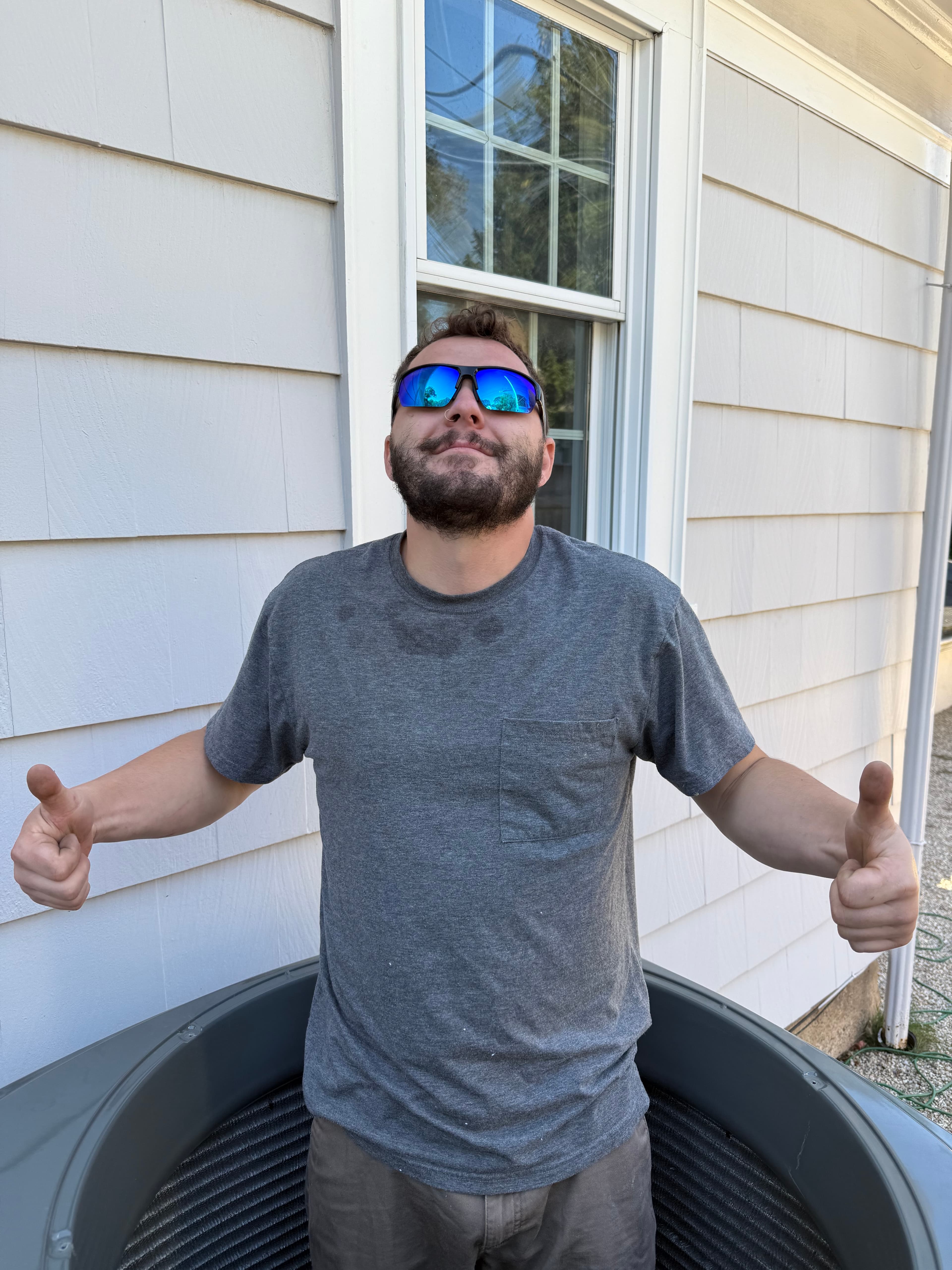 Bearded man in blue sunglasses gives two thumbs up while standing in an AC unit.